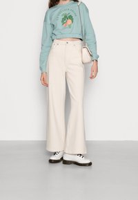Pantalones de campana de color claro hechos de tela texturizada, combinados con un sudadera corta de color verde menta con un diseño gráfico, y zapatos blancos.