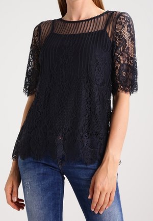 Žena v tmavě modré krajkové blůze s průsvitným pruhovaným sedlem a krátkými rukávy, doplněná modrými džínami z denimu.