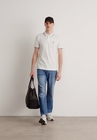 Biela polo tričko s čiernym lemovaním, modré džínsy a šedé tenisky. Nesie čiernu tašku. Model má na hlave čiernu čiapku. Hladký, neformálny outfit.