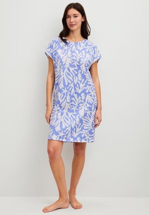 Frau steht barfuß und trägt ein knielanges blaues Kleid mit weißen Korallen- und Meeresbewohner-Mustern vor schlichtem hellem Hintergrund.