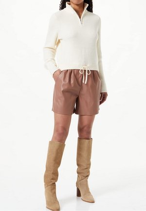 Frau trägt einen cremefarbenen Reißverschluss-Pullover, braune Leder-Shorts mit Taillenband und beige Wildleder-Kniehohe Stiefel mit Absatz, steht mit den Händen in den Taschen.
