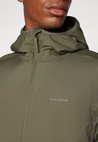 Maslinasto zelena zip-up jakna s visokim ovratnikom s kapuljačom, teksturirana tkanina i suptilan "SALOMON" logotip u srebrnoj boji na prsima.