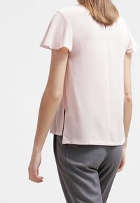 Ljusrosa blus med korta ärmar, med lätt volang på ärmarna, rak skärning och sidoplikter i fållen. Slät textur och lättviktig tyg.