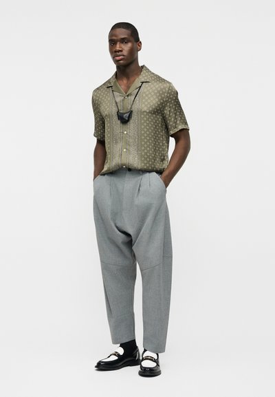 Άνδρας που φοράει πουκάμισο με κοντά μανίκια σε ελιά με σχέδιο, γκρι ψηλόμεσα παντελόνια με πιέτες, μοκασίνια ασπρόμαυρα και μαύρη μάσκα προσώπου γύρω από το λαιμό.