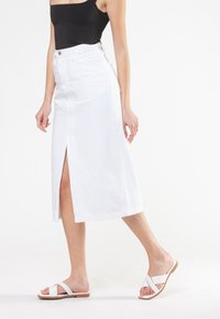Hvid denim midi-nederdel med en frontslids, høj talje og fem-lomme design. Parret med flade, hvide sandaler med et minimalistisk strop.