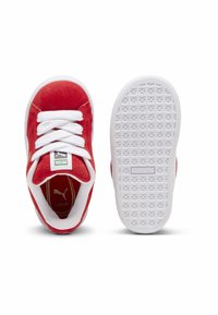 Adidași roșii din suede cu dungi albe și accente. Prezintă o talpă texturată din cauciuc alb și un detaliu cu logo de aur în interior.
