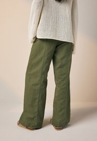 Pantaloni largi, de culoare verde măsliniu, confecționați dintr-un material ușor, cu cusături frontale și croi lejer. Asortați cu un pulover crem tricotat.