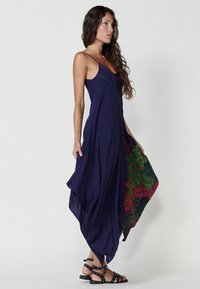 Robe de marine avec un design fluide, ornée de clous décoratifs et de motifs floraux vifs en rouge et vert le long de l'ourlet. Sandales à lanières portées.