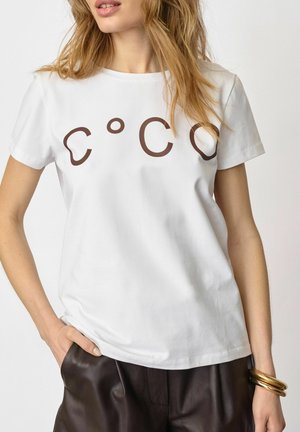 Kobieta w białej koszulce z brązowym abstrakcyjnym napisem „COCO”, ciemnobrązowe spodnie i złote bransoletki na prawej dłoni, ręka w kieszeni.