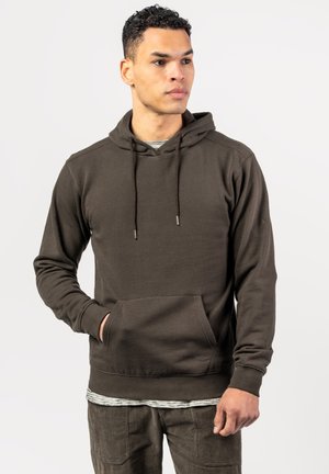 Young man with curly hair wearing a dark brown hoodie and matching pants, looking slightly to the side with one hand in the hoodie pocket.