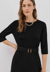 Czarna sukienka do kolan z rękawami o długości trzech ćwiartek, z dopasowaną talią i paskiem z prostokątną złotą klamrą. Gładka tekstura, minimalistyczny design.