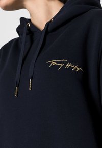 Navyblauwe hoodie gemaakt van zachte stof, voorzien van een gouden geborduurd logo op de borst en gouden accenten op de verstelbare koorden.