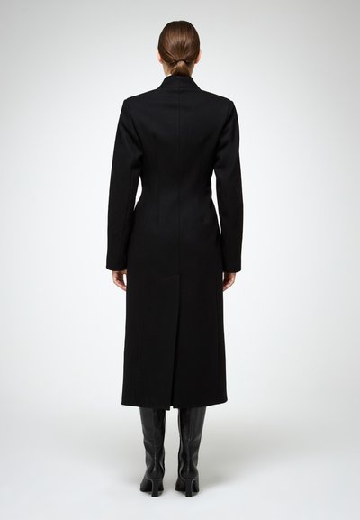 Lungo cappotto nero in lana, con silhouette aderente, maniche lunghe, spacco posteriore e colletto a punte. Indossato con stivali neri al ginocchio.