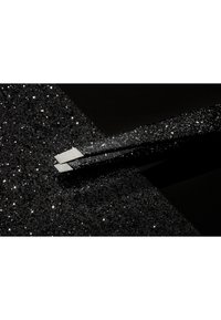 Pince à épiler noire avec une finition brillante, dotée d'une prise texturée ressemblant à des paillettes, posée sur une surface noire scintillante.