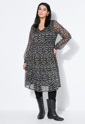 Kvinde med mørkt hår iført en gennemsigtig kjole med leopardprint, sorte leggings og sorte cowboystøvler, stående mod en hvid baggrund.