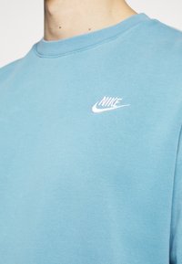 Světlé modrá mikina z měkkého materiálu s bílým vyšívaným logem Nike na levé straně hrudi. Klasický kulatý výstřih a jednoduchý design.