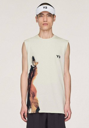 Junger Mann, der ein ärmelloses beiges Oberteil mit abstraktem braun-schwarzem Muster, schwarze Hosen und ein weißes Y-3-Visier trägt, steht vor einem einfarbigen Hintergrund.