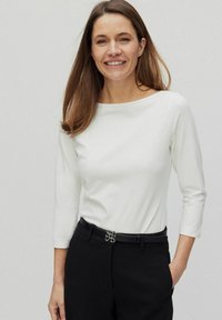 Haut à manches longues blanc avec un décolleté bateau associé à un pantalon noir. Le pantalon est orné d'une boucle de ceinture en argent décorative.