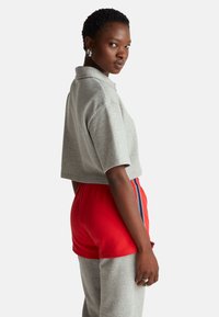 Šedé zkrácené polo tričko s krátkými rukávy, spojené s červenými šortkami s bočními pruhy. Látka vypadá měkce a volně.