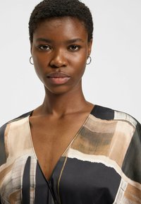 Blouse à motifs abstraits en tissu doux, présentant un décolleté en V profond, des tons bruns et noirs, et des accents clairs. Pas de matériel visible.