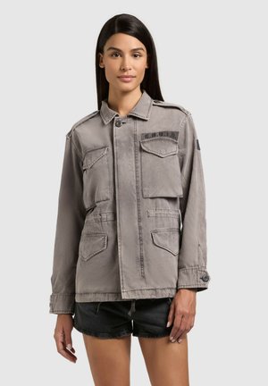 Woman wearing a gray utility jacket with multiple pockets and black shorts, standing against a plain light background.