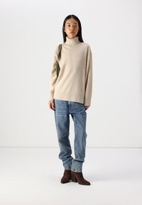 Un pull col roulé beige en tricot associé à un jean léger, ample et bleu, ainsi qu'à des bottines marron à talons carrés.