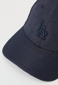 Boné azul-marinho feito de tecido acolchoado com costura hexagonal e um logo bordado "LA" em azul correspondente. Inclui uma aba curva.