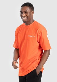 Usměvavý muž v jasně oranžovém tričku s nápisem "SMILODOX Ltd. ", stojící s jednou rukou v kapse před jednobarevným pozadím.