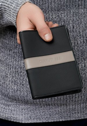 Roka drži črni usnjeni denarnici z bež progo in vgraviranim napisom "CERRUTI 1881" na ozadju sive pletene majice.