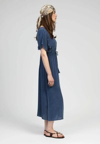 Robe bleue, longueur cheville, avec des manches courtes volantes, une taille à cordon et des fentes sur les côtés ; portée avec un foulard à motifs et des sandales plates.