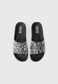 Sandales noires à glissière avec un semelle texturée et une large sangle ornée du texte contrastant "Versace Jeans Couture" en lettres majuscules.