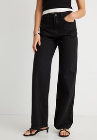 Frau trägt hoch taillierte, weit geschnittene schwarze Jeans, schwarze Riemchen-Sandalen mit niedrigem Absatz und ein schwarzes kurzes Oberteil, eine Hand in der Tasche.