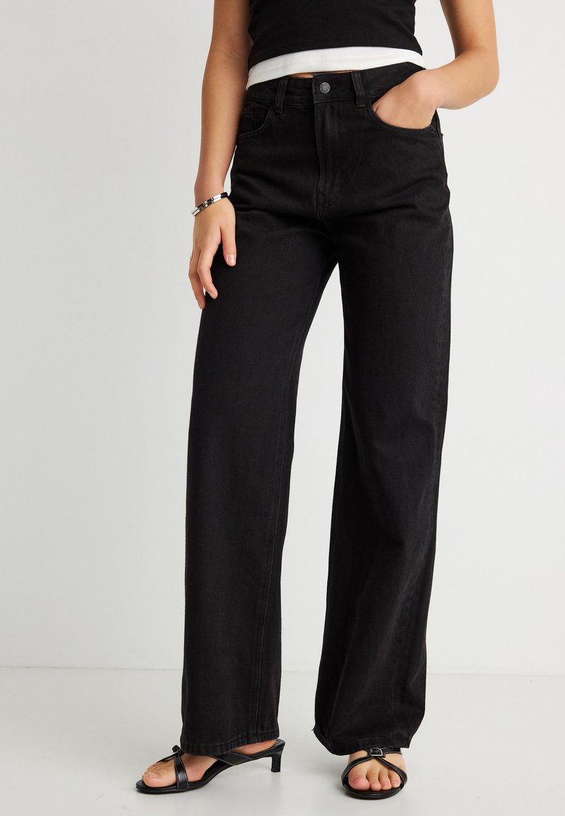 Frau trägt hoch taillierte, weit geschnittene schwarze Jeans, schwarze Riemchen-Sandalen mit niedrigem Absatz und ein schwarzes kurzes Oberteil, eine Hand in der Tasche.