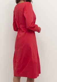 Rode jurk met lange mouwen, een aansluitende bovenkant en een uitlopende rok. Gemaakt van soepele stof, met een ronde halslijn en een knoopsluiting aan de achterkant.