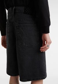 Pantalones cortos de mezclilla oscura con corte recto, dos bolsillos traseros y una etiqueta de cintura a cuadros. La tela tiene una textura suave.