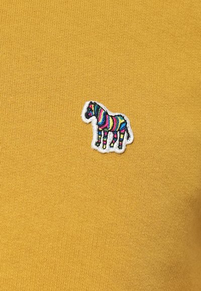 Κίτρινο πλεκτό ύφασμα με μια μικρή κεντημένη ετικέτα ζέβρας σε ζωντανά χρώματα, περιγραμμισμένη με λευκές ραφές, τοποθετημένη από αριστερά.