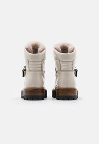 Bottines en cuir verni blanc avec doublure en fausse fourrure, talons en bois, et détail de boucle sur le côté. Surface texturée et accents cousus.