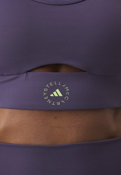 Lilla sportsbh med utskjæringsdesign. Har en sirkulær logo i neongrønt med merkenavn og tre striper i bunnen. Glatt tekstur.