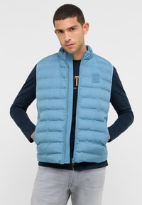 Mann in einer hellblauen, gesteppte ärmellosen Weste über einem schwarzen Langarmshirt, die Hände in den Taschen der Weste, stehend vor einem weißen Hintergrund.