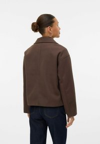 Brūna pārmēra jaka ar atšķeltu apkakli, nolaižamām pleciem un teksturētu audumu, redzama no aizmugures kopā ar zilajām džinsa biksēm.