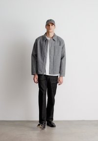 Grau gewebte Jacke mit Knopfleiste, Brusttasche, weißem T-Shirt, dunklen Hosen und grauem Cap. Glatte Textur und lässige Passform.