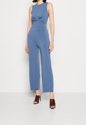 Femme portant une combinaison bleue sans manches avec un détail torsadé sur le devant et des mules à talons hauts ouvertes marron, debout devant un fond blanc uni.