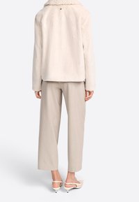 Femme portant une veste en fausse fourrure beige clair, un pantalon cropped large beige et des escarpins à talons kitten crème, se tenant de dos.