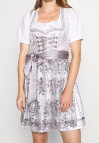 Eine Frau trägt ein silbernes und lavendelfarbenes Dirndl mit floraler Stickerei, einem mit einer Schleife gebundenen Satin-Tailleband und einer weißen Puffärmel-Bluse.