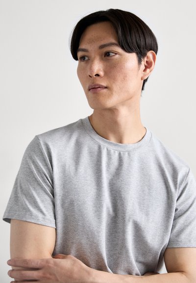 T-shirt en coton gris clair avec un col rond. Présente des manches courtes et une texture lisse, présentée sur une personne les bras croisés.