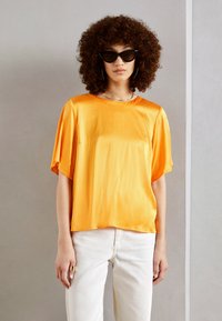 Top in satin arancione brillante con maniche corte e vestibilità comoda, abbinato a pantaloni bianchi. La trama è liscia, con un semplice scollo rotondo.