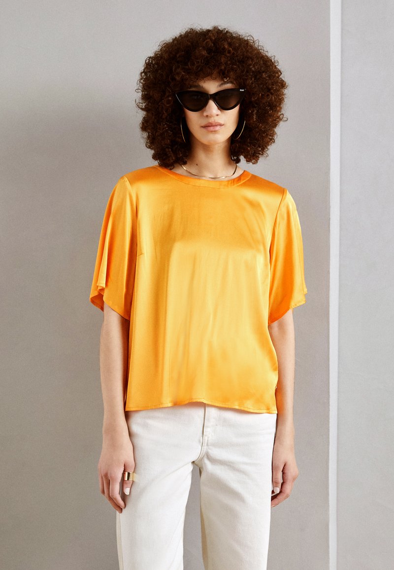 Top in satin arancione brillante con maniche corte e vestibilità comoda, abbinato a pantaloni bianchi. La trama è liscia, con un semplice scollo rotondo.