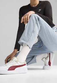 Baskets blanches avec des accents rouges et beiges, dotées d'un dessus en cuir texturé, d'un système de laçage et d'une semelle en caoutchouc. Portées avec un jean bleu clair.