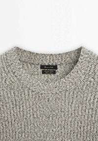 Col rond en grosse maille grise avec col côtelé et étiquette noire indiquant la marque et les détails du tissu.