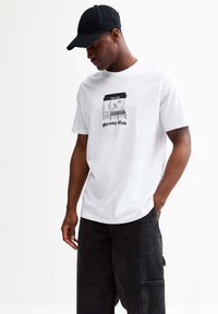 Camiseta de algodón blanca con un gráfico negro de una cafetería y el texto "Morning Club" debajo, combinada con pantalones cargo oscuros.
